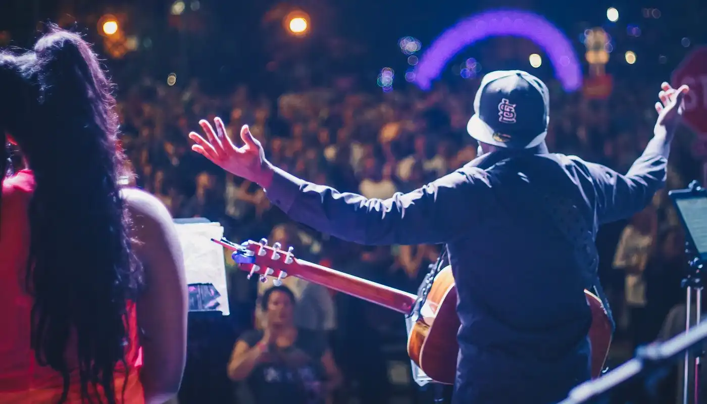 A man holds a guitar from a behind on stage with a crowd before him