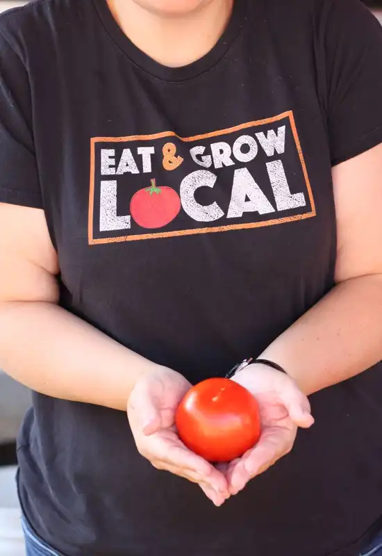 A person holds a fresh tomato with a shirt that reads "Eat & Grow Local"