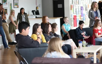 A group of young adults sit and stand in a room with attention directed towards a speaker