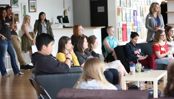 A group of young adults sit and stand in a room with attention directed towards a speaker