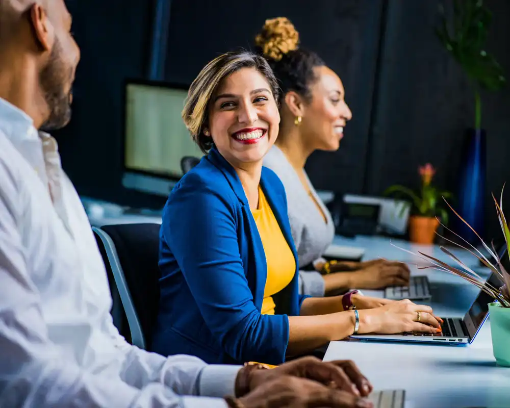 Three people sit at computers and smile towards one another