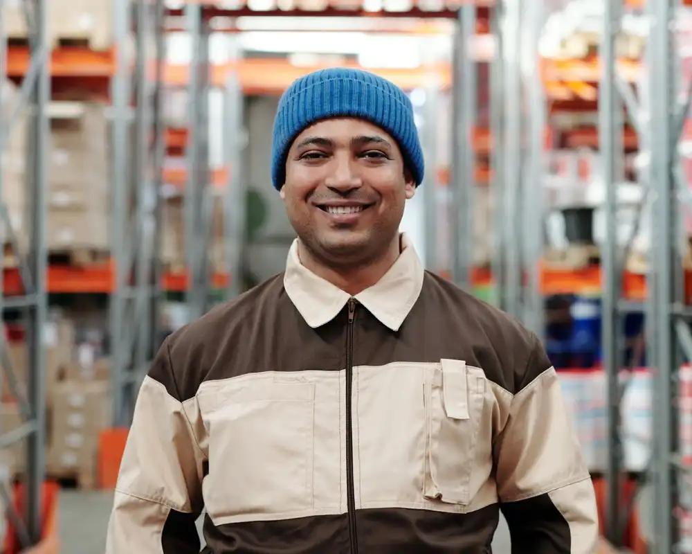 A man in a blue beanie smiles at the camera while standing in a large warehouse with shelves